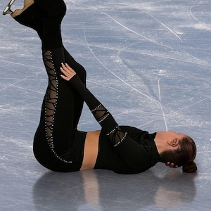 Könnte beinhalten: Eine Eiskunstläuferin in schwarzem Outfit führt eine Bewegung auf dem Eis aus. Die Läuferin ist kopfüber mit ausgestreckten Beinen, trägt schwarze Leggings mit Schnürdetails. Die Kufen der Eislaufschuhe sind sichtbar.