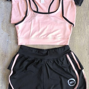 Könnte beinhalten: Ein dreiteiliges Sportoutfit in Schwarz und Rosa. Das Outfit besteht aus einem schwarz-rosa T-Shirt mit kurzen Ärmeln und Logo, einem rosa Sport-BH mit schwarzem Besatz und schwarzen Sport-Shorts mit rosa Besatz und Logo.