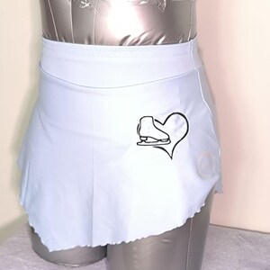 May include: A pale blue ice skating skirt featuring a black figure skate and heart design. The skirt has a scalloped edge and is shown on a mannequin. This skirt is designed for ice skating and is a performance garment.
