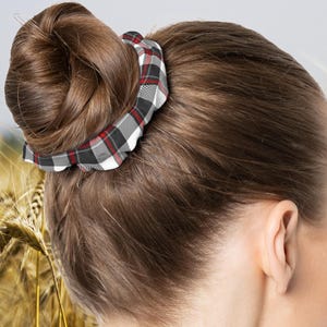 Op de afbeelding: Een knotkapsel vastgezet met een geruite scrunchie. De scrunchie heeft een rood, zwart en wit ruitpatroon. De achtergrond toont een tarweveld. Een vrouw is van achteren te zien.