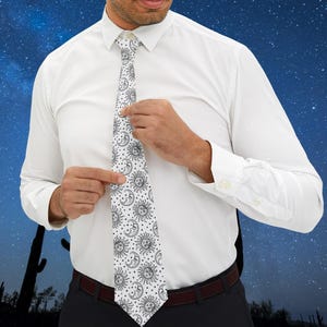 Può includere: Una camicia bianca con una cravatta bianca e nera con un motivo ripetitivo di soli e lune. La cravatta viene regolata da una persona che indossa pantaloni neri.