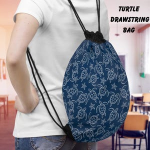 Puede incluir: Una bolsa con cordón de color azul marino con un estampado de tortugas y estrellas de mar blancas. La bolsa tiene cordones negros y un cierre negro. El texto "TURTLE DRAWSTRING BAG" está impreso en blanco en la parte superior derecha.