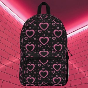 May include: Black backpack with a pink heart pattern. The hearts are outlined in a darker pink and have a gradient effect.