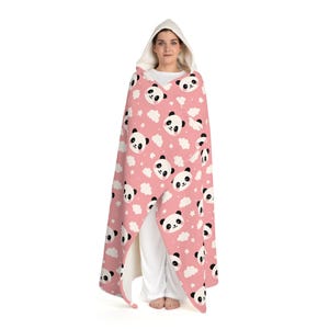 Puede incluir: Una manta con capucha rosa con un estampado de caras de panda, nubes blancas y estrellas. La manta tiene un interior blanco. La persona lleva la manta.