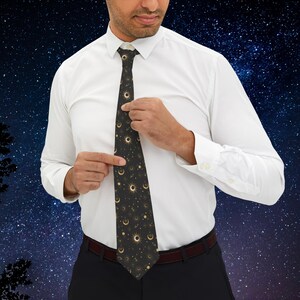 Puede incluir: Una corbata negra con un patrón celestial dorado con lunas, estrellas y soles. La corbata la lleva puesta una persona sobre un fondo azul oscuro con estrellas blancas.