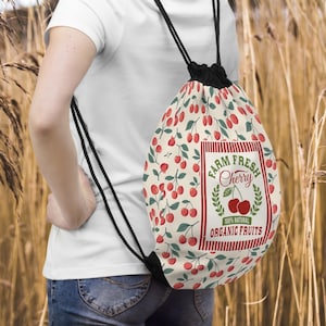 May include: A drawstring backpack with a cream background covered in red cherries and green leaves. The bag features a rectangular label with the words "FARM FRESH Cherry" and "100% NATURAL ORGANIC FRUITS". The bag has black drawstrings.