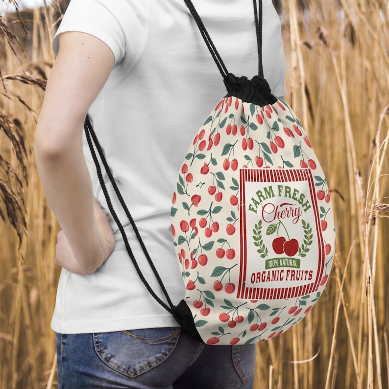 May include: A drawstring backpack with a cream background covered in red cherries and green leaves. The bag features a rectangular label with the words "FARM FRESH Cherry" and "100% NATURAL ORGANIC FRUITS". The bag has black drawstrings.