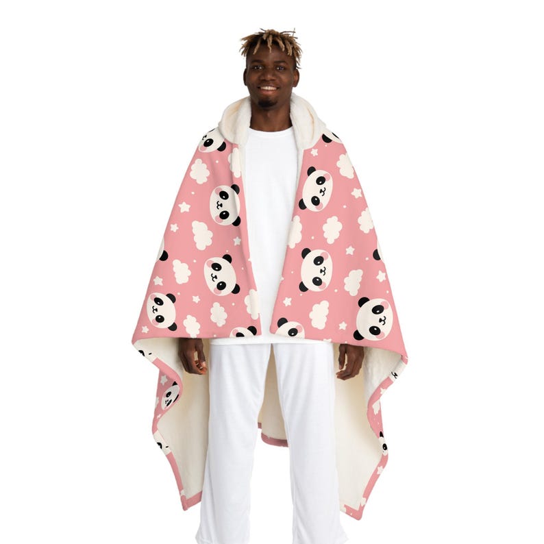 Puede incluir: Una manta con capucha con un estampado de pandas rosas, nubes blancas y estrellas. La manta tiene un forro de sherpa blanco y un ribete rosa. La persona lleva una camiseta blanca y pantalones blancos.