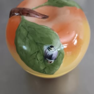 May include: A ceramic apple-shaped container with a lid. The container is painted in shades of orange and yellow, with green leaves and a brown stem. A small, decorative bug is on one of the leaves. The container is likely used for storage.