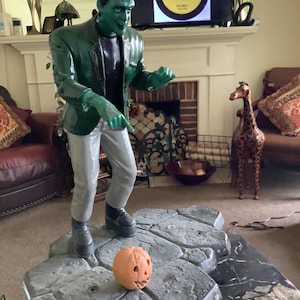May include: A large Frankenstein's monster statue with green skin, a black jacket, and gray pants. It stands on a rocky base next to a carved pumpkin. The statue is a Halloween decoration. A television in the background displays a record album.