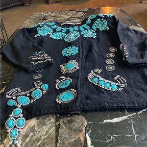 May include: Black cardigan sweater adorned with turquoise-colored stones and silver beadwork. The sweater features intricate floral and geometric designs, with embellishments on the front, sleeves, and neckline. A unique, eye-catching piece.