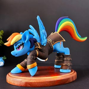 Könnte beinhalten: Blaue Ponyfigur mit Regenbogen-Schweif und Flügeln, in einem braunen Outfit mit goldenen Akzenten. Die Figur steht auf einem Holzsockel mit detailliertem Design und leuchtenden Farben.