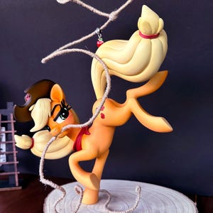 May include: A cartoon pony figurine in a dynamic pose, made from smooth, light orange material. The pony has a brown hat, red bow, and a rope lasso. The figurine is mounted on a wooden slice base, set against a dark background.