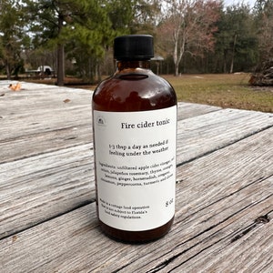May include: A brown glass bottle of "Fire cider tonic" with a black cap. The label lists ingredients like apple cider vinegar, onion, and ginger. The bottle is 8 oz and is on a weathered wooden surface, with a blurred outdoor background.