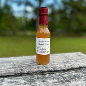 May include: A small glass bottle with a red cap and a white label. The label reads "Crenshaw Farms Almond extract made with bitter apricot kernels and American vodka 5 oz".