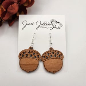 May include: A pair of wooden acorn earrings with a delicate, intricate design. The earrings are brown and have a silver hook.