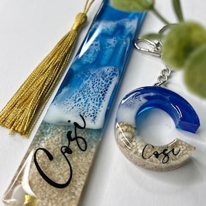 Könnte beinhalten: Ein Lesezeichen- und Schlüsselanhänger-Set aus Kunstharz mit Strandmotiv. Das Lesezeichen hat ein blau-weißes Wellenmuster mit einer goldenen Quaste. Der Schlüsselanhänger ist ein blau-weißer Buchstabe "C" mit Sand und Muscheln. Der Name "Cosi" steht auf beiden Artikeln.