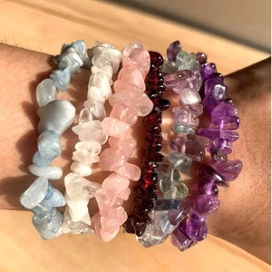 Pode incluir: Um conjunto de sete pulseiras de pedras preciosas, cada uma com uma cor e tipo de pedra diferente. As pulseiras são feitas de pequenas pedras de forma irregular e estão empilhadas no pulso de uma pessoa. As cores incluem azul claro, branco, rosa, vermelho, verde e roxo.