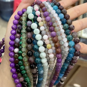 Pode incluir: Uma coleção de 14 fios de contas de pedras preciosas, cada fio com uma cor e tipo de pedra diferente. As contas são redondas e lisas, e as cores incluem roxo, verde, azul, marrom e branco.