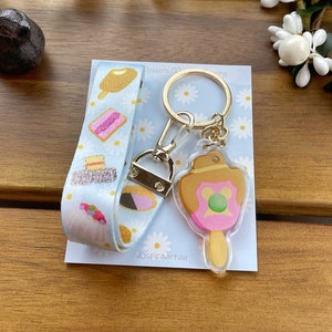 May include: A blue and white ribbon with a variety of desserts printed on it is attached to a gold key chain with a clear charm featuring a cartoon popsicle with a pink, green, and brown design. The charm is labelled "Dspicaarttau".