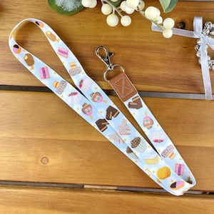 Puede incluir: Un cordón azul y blanco con un llavero de cuero marrón. El cordón presenta un patrón de dulces y golosinas australianas, incluyendo Tim Tams, Cobbers Candy y Fairy Bread.