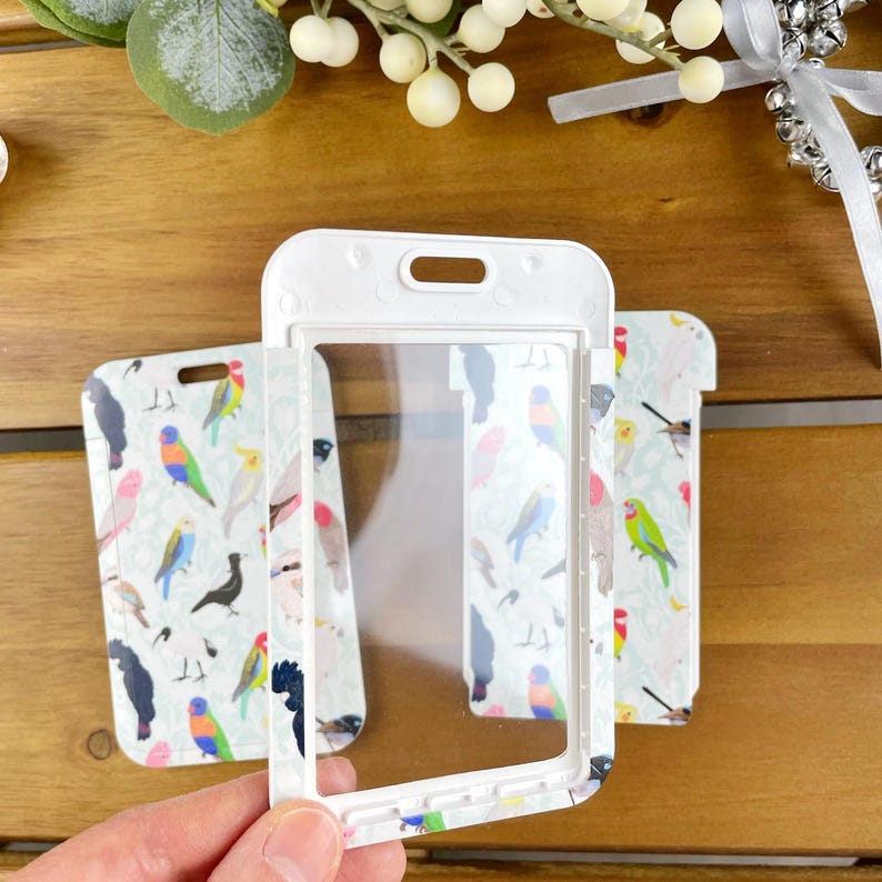 May include: A white plastic vertical badge holder with a clear rectangular window. The frame features a repeating pattern of colorful birds including parrots, cockatoos, and other avian species in shades of green, blue, pink, and black.