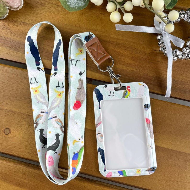 May include: A light green lanyard and matching vertical ID badge holder featuring a repeating pattern of various colorful Australian birds including cockatoos and kookaburras. The set includes a brown faux leather connector and a silver metal clasp.
