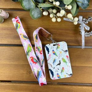 May include: A light pink lanyard and matching vertical ID badge holder featuring a colorful pattern of various parrots and white daisies. The lanyard includes a brown faux leather connector and a silver metal swivel hook attached to the badge holder.