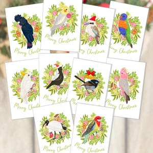 May include: A collection of ten white greeting cards featuring watercolour illustrations of various birds, including cockatoos, a kookaburra, and a magpie, surrounded by green wreaths with pink flowers and the text Merry Christmas in gold script.