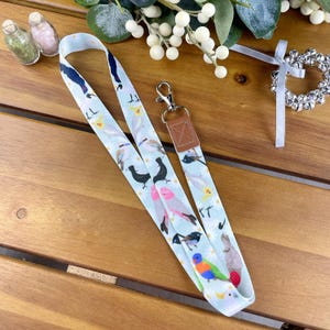 May include: A white lanyard with a colourful bird pattern and a brown leather key ring. The lanyard has a silver metal clip.