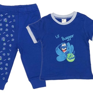 Può includere: Un completo per neonato in due pezzi blu e azzurro. La maglietta presenta un personaggio dei cartoni animati con il testo "Lil' Bugger". I pantaloni hanno un motivo ripetuto di un personaggio dei cartoni animati.