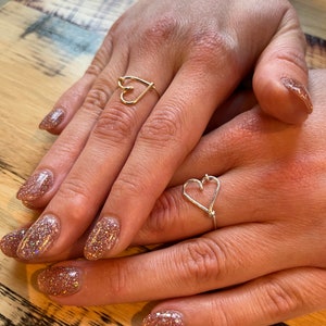 May include: Two hands with gold glitter nail polish wearing two heart-shaped rings. One ring is gold and the other is silver.