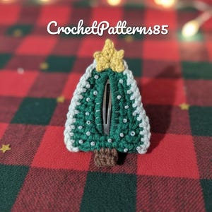 May include: A handmade green and white crochet Christmas tree hair clip with a gold star on top. The hair clip is decorated with small white beads and has the text "CrochetPatterns85".