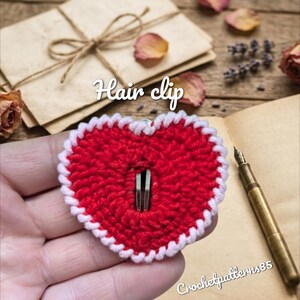 May include: A red heart-shaped hair clip with a white border. The clip is made of a crocheted material. The text "Hair clip" is written in white script above the clip. The text "Crochetpatterns85" is written in white script below the clip.
