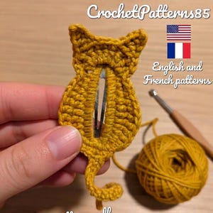 May include: A mustard yellow crocheted cat-shaped hair clip with a silver metal snap barrette embedded in the centre. The design features pointed ears and a curled tail. Text on the image reads CrochetPatterns85, English and French patterns.