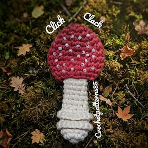 May include: A handmade crochet mushroom with a red cap dotted with white beads and a white stem. The mushroom is set against a mossy background with fallen leaves. Text on the image says "Click Clack" and "Crochetpatterns85".