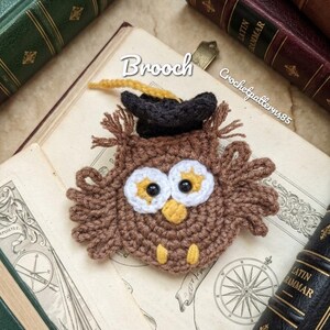 May include: A handmade owl brooch, crocheted in brown yarn with white and yellow details, wearing a black graduation cap with a yellow tassel. The image includes the text "Brooch" and "Crochetpatterns85".