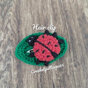 Può includere: Un fermaglio per capelli fatto a mano all'uncinetto con una coccinella rossa con pois e dettagli neri, posto su una base verde a forma di foglia. I testi "Hair clip" e "Crochetpatterns85" sono visibili.