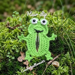May include: A green crocheted frog hair clip with white and black eyes, a black smile, and a silver clip. The text "Hair clip" is above the frog, and "Crochetpatterns85" is below. The clip is set against a background of green moss.
