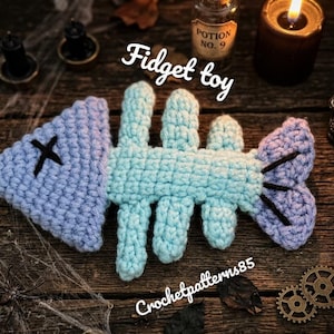 May include: A handmade crochet fidget toy shaped like a fish skeleton, in shades of blue and teal, with a black cross and black accents. The text "Fidget toy" is visible. The background includes a candle, skull, and gears.