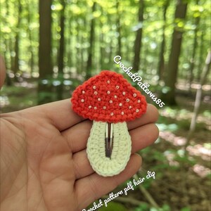 May include: A handmade crochet mushroom hair clip. The mushroom cap is red with white dots, and the stem is cream-colored. The text "CrochetPatterns85" and "Crochet pattern pdf hair clip" are visible. The clip is held against a blurred forest background.