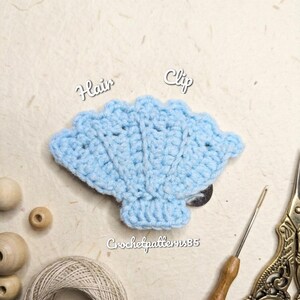 May include: A light blue, crocheted seashell hair clip. The handmade clip features a detailed, textured design and is displayed with craft supplies, including a crochet hook, scissors, and the text "Hair Clip". The text "Crochetpatterns35" is also visible.