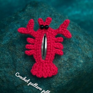 May include: A handmade red crochet lobster hair clip with black bead eyes and a silver barrette clip. The text "CrochetPatterns85" and "Crochet pattern pdf" are written in white cursive. The clip is set against a blue-green stone.