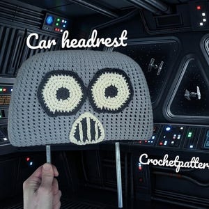May include: A grey, crocheted car headrest cover with large, circular eyes and a mouth-like design. The headrest has a whimsical, cartoonish appearance. The text "Car headrest" is above the headrest, and "Crochetpatterns85" is below.