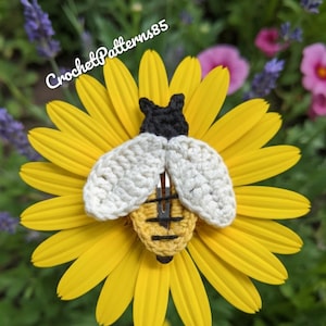 May include: A handmade crochet bee hair clip with white wings, a yellow body, and a black head, resting on a bright yellow flower. The text "CrochetPatterns85" is visible in the image. The background features green foliage and other flowers.