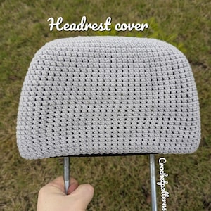 May include: A light grey, crocheted headrest cover. The cover is rectangular with rounded edges and is held by two metal posts. The text "Headrest cover" is written in cursive across the top of the image.