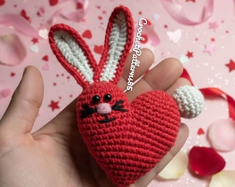Könnte beinhalten: Ein handgehäkelter roter Hase mit herzförmigem Körper. Der Hase hat weiß gefütterte Ohren, eine kleine rosa Nase und einen weißen Bommel-Schwanz. Der Text "CrochetPatterns85" ist sichtbar. Der Hintergrund zeigt rosa und rote Herzformen und Bänder.