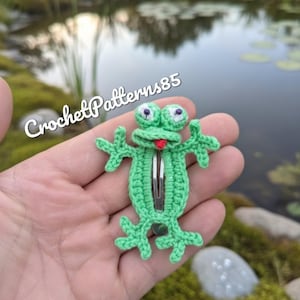 Puede incluir: Una pinza para el pelo de rana verde tejida a ganchillo, con una lengua roja y ojos saltones. La pinza para el pelo está hecha de hilo verde y tiene el texto "CrochetPatterns85" en blanco.