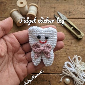 May include: A handmade crochet fidget clicker toy shaped like a tooth, with a pink bow and smiling face. The toy is white with black eyes and a red smile. The text "Fidget clicker toy" is above the toy.