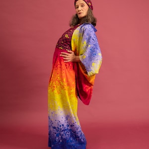 K&ouml;nnte beinhalten: Eine Person tr&auml;gt ein farbenfrohes Outfit mit einem Regenbogen-Ombre-Muster. Das Outfit besteht aus einem lang&auml;rmeligen Oberteil mit Sternenmuster, einer Hose und einem Turban. Die Person steht vor einem rosa Hintergrund.